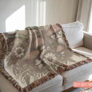 May include: A cozy sofa throw featuring a whimsical design of mushrooms, leaves, and flowers in shades of brown, green, and white. The throw has a fringed edge and is made of a soft, woven fabric.