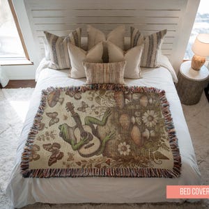 May include: A white bed with a green frog playing a guitar on a brown and green tapestry bed cover. The bed cover has a fringe border and features butterflies, flowers, and mushrooms.
