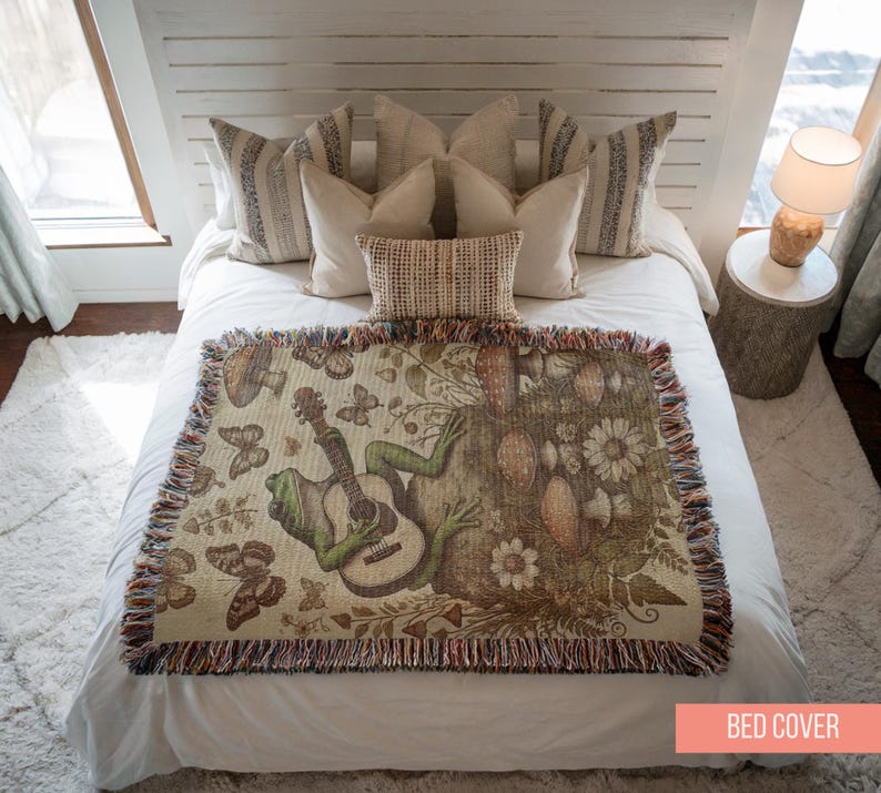 May include: A white bed with a green frog playing a guitar on a brown and green tapestry bed cover. The bed cover has a fringe border and features butterflies, flowers, and mushrooms.