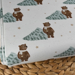 May include: White fabric with a repeating pattern of brown bears wearing white scarves, green trees, and gold stars. The fabric is folded, showing the pattern's repeat. The fabric is likely for sewing or crafting.