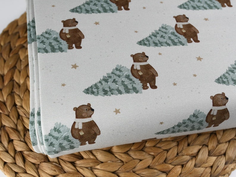 May include: White fabric with a repeating pattern of brown bears wearing white scarves, green trees, and gold stars. The fabric is folded, showing the pattern's repeat. The fabric is likely for sewing or crafting.