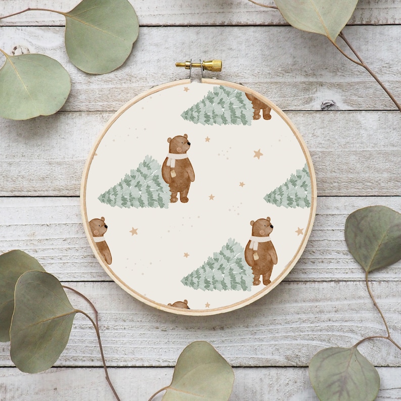 May include: A round embroidery hoop featuring a repeating pattern of brown bears wearing white scarves, carrying green Christmas trees. The background is a cream color with small gold stars. The hoop is made of wood.