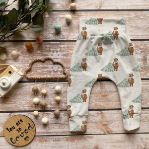 May include: Cream-colored baby pants with a repeating pattern of brown bears wearing scarves and green trees. The pants have a high waistband and cuffed ankles. The words "You are so Loved" are on a wooden plaque.