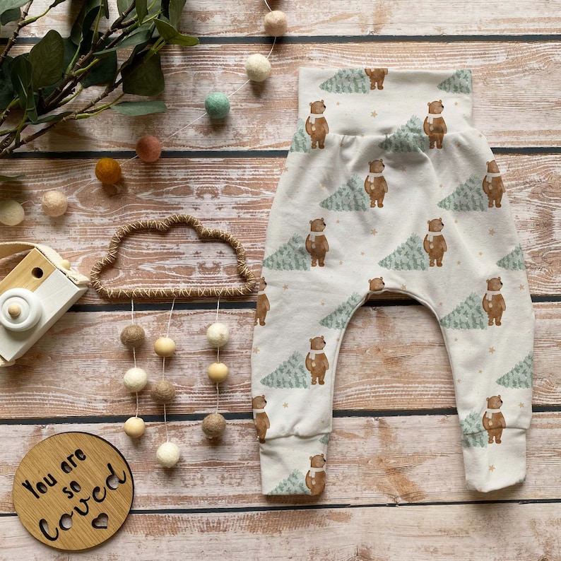 May include: Cream-colored baby pants with a repeating pattern of brown bears wearing scarves and green trees. The pants have a high waistband and cuffed ankles. The words "You are so Loved" are on a wooden plaque.