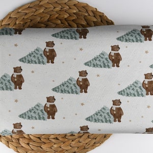 May include: A fabric with a repeating pattern of brown bears wearing white scarves next to green Christmas trees. Small tan stars are scattered across the off-white background. The fabric is folded and resting on a woven wicker surface.
