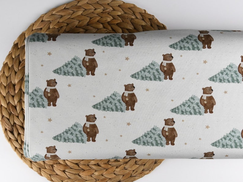 May include: A fabric with a repeating pattern of brown bears wearing white scarves next to green Christmas trees. Small tan stars are scattered across the off-white background. The fabric is folded and resting on a woven wicker surface.