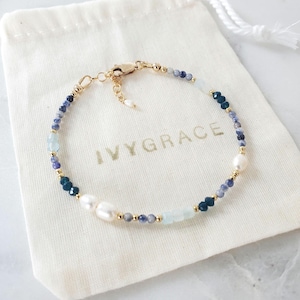Peut inclure: Un bracelet doré délicat orné de perles bleues, blanches et nacrées. Le bracelet est posé sur un sac en tissu blanc avec le texte "IVYGRACE" en lettres dorées. Le bracelet a un fermoir doré.