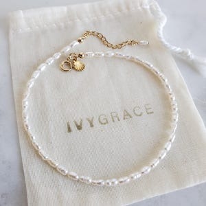 May include: A delicate pearl bracelet with a gold chain and seashell charm rests on a cream-coloured pouch. The pouch is printed with the text "IVYGRACE" in gold. The bracelet is made of small, oval-shaped pearls.