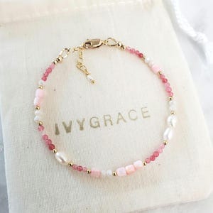 May include: A delicate bracelet with pink and white beads, small gold beads, and freshwater pearls. The bracelet is displayed on a white cloth bag with the text "IVYGRACE" printed in gold.