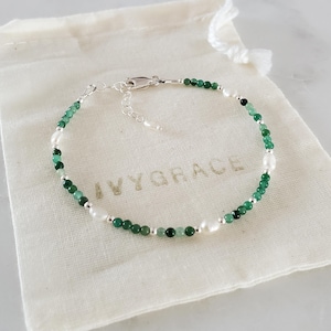 Peut inclure: Un bracelet délicat composé de petites perles rondes vert émeraude et de perles blanches ovales. Le bracelet est monté sur une chaîne argentée et repose sur une pochette crème avec le texte "IVYGRACE".