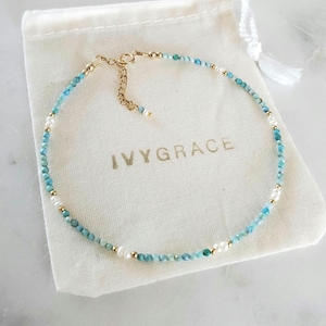 Könnte beinhalten: Eine zarte Halskette mit kleinen, hellblauen und weißen Perlen, die mit goldfarbenen Akzenten durchsetzt sind. Die Halskette wird auf einem weißen Kordelzugbeutel mit dem Aufdruck "IVYGRACE" präsentiert.