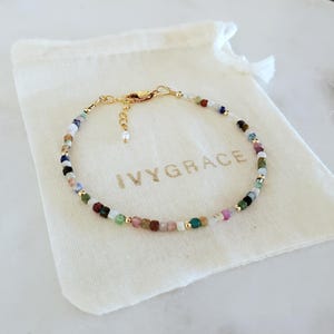 Op de afbeelding: Een delicate armband gemaakt met een verscheidenheid aan gekleurde edelsteen kralen. De armband ligt op een witte stoffen tas met de tekst "IVY GRACE" in goud gedrukt.