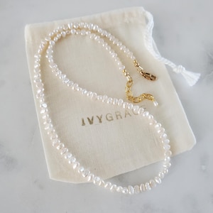 May include: A delicate pearl necklace with a gold clasp and extender chain rests on a cream-coloured pouch. The necklace features small, irregularly shaped white pearls. The pouch has the text "IVYGRACE" printed on it.