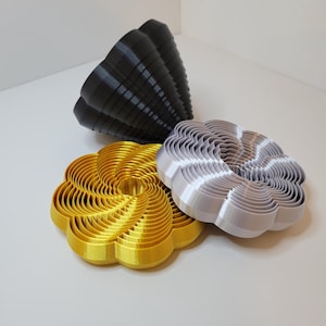 Puede incluir: Tres cortadores de galletas impresos en 3D con forma de flores. Los cortadores están en capas y tienen un diseño en espiral. Los cortadores son dorados, plateados y negros.