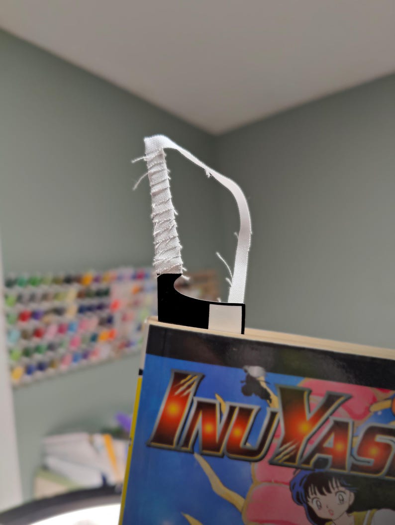 May include: A bookmark with a white fabric loop and a black and white rectangular base. The bookmark is inserted into a book with the title "InuYasha" in gold and red lettering on a blue background.