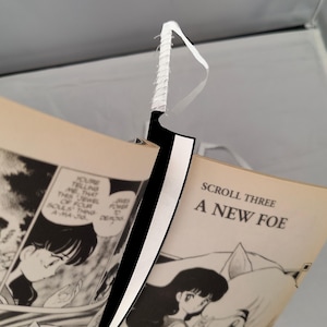 May include: A black and white decorative sword with a white handle is placed between the pages of a manga comic book. The comic book pages are open, displaying black and white illustrations and text. The text on the right page reads "SCROLL THREE A NEW FOE."
