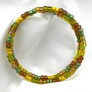May include: A multi-strand beaded bracelet with green, yellow, and orange beads. The bracelet is made of small, round beads that are strung together in a repeating pattern.