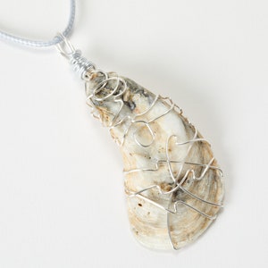 May include: A pendant necklace featuring a natural oyster shell wrapped in silver-colored wire. The shell has a cream and tan color with a unique pattern. The necklace hangs from a light gray cord.