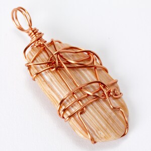 May include: A copper wire wrapped pendant featuring a light brown, striated stone. The wire is wrapped in a decorative pattern around the stone.