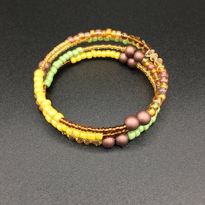 May include: A beaded wrap bracelet featuring yellow, green, and gold beads. The bracelet has a spiral design with brown accent beads. The beads are arranged in rows, creating a textured and colorful accessory.