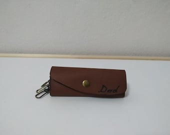 May include: A brown leather key holder with a brass button and a key ring. The key holder is personalized with the word "Dad" in black lettering.