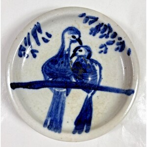 Pode incluir: Um pequeno prato redondo de cerâmica com um desenho pintado à mão de dois pássaros azuis empoleirados num galho. As aves são retratadas num estilo simples, de arte popular, sobre um fundo branco. Folhas decorativas circundam os pássaros.