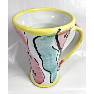 May include: A ceramic mug with a white interior, yellow rim and handle. The exterior has abstract designs in pink, teal, and black against a white background. The mug has a flared shape.