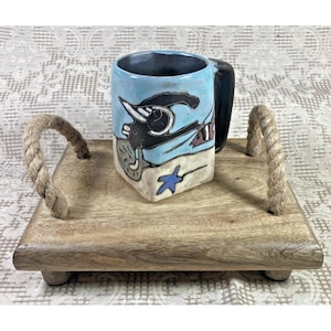 May include: A wooden serving tray with rope handles, featuring a blue and white ceramic mug with a whimsical sea creature design. The mug has a black handle and a blue glaze.