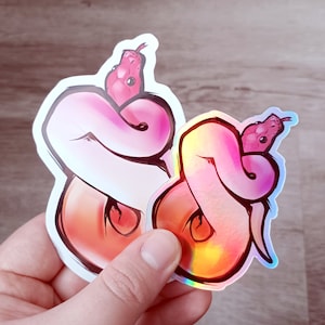 May include: Two iridescent stickers featuring a pink and red snake curled up in a circle. The stickers are on a light brown wooden surface.