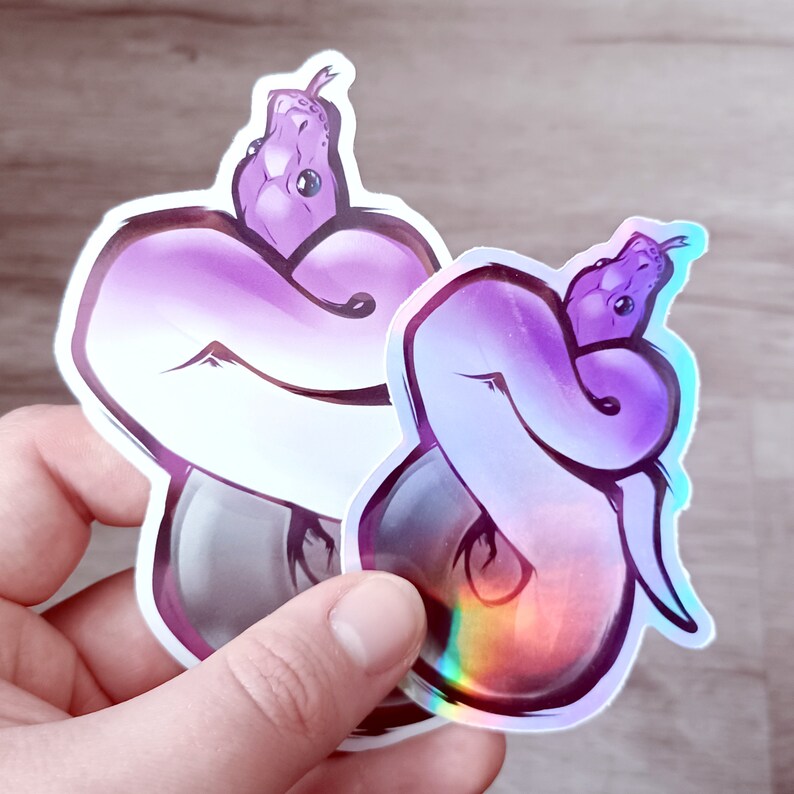 May include: Two iridescent stickers of a purple snake curled up. The sticker on the left is on a white background, while the sticker on the right is on a holographic background.
