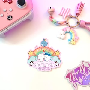 Puede incluir: Un mando de videojuegos rosa está junto a una pegatina con el texto "Let's Take Ibuprofen Together" sobre un arcoíris y personajes de dibujos animados. También hay un llavero con un arcoíris, una estrella y una borla.