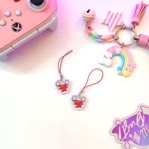 May include: Two clear acrylic charms with a red cartoon character and red strings. A pink game controller is in the background. A charm bracelet with a rainbow, star, and bell is also visible.