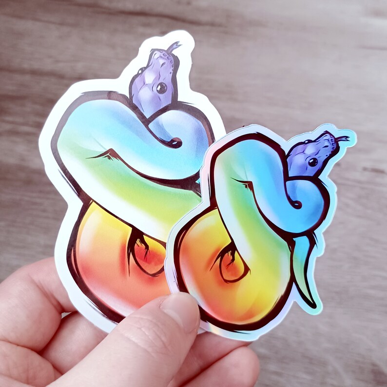 May include: Two rainbow colored snake stickers with purple snake heads. The stickers are on a wood background.