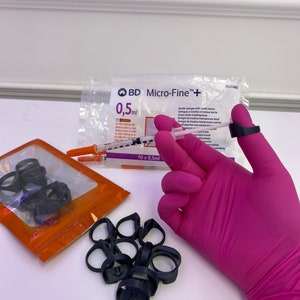 May include: A close-up of a pink latex glove holding a syringe with a needle. The syringe is labeled "BD Micro-Fine + 0,5ml". There are also black rubber rings and a clear plastic bag with a syringe and a label that says "10 x 0,5ml".