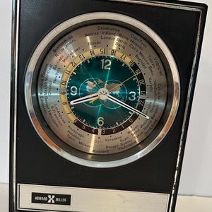 May include: A rectangular black Howard Miller world time clock with a circular silver face. The dial features a green map, white numerals at 3, 6, 9, and 12, and a rotating outer ring listing global cities and a 24-hour time scale.