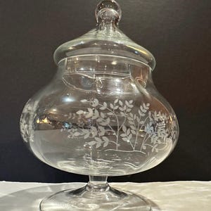May include: A clear glass candy dish with a matching lid, featuring a pedestal base and a wide, rounded body. The exterior of the glass is decorated with a delicate, etched white floral pattern depicting leafy branches and small blossoms.