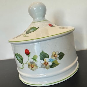 May include: A white ceramic jar with a lid, decorated with a strawberry and floral design. The jar has a rounded base and a lid with a knob. Green trim accents the edges of the jar and lid.