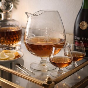 May include: A clear glass decanter and brandy snifters filled with amber liquid sit on a mirrored surface. A bottle of Remy Martin cognac is in the background. A plate of lemon slices and ice tongs are also present.