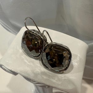 Peut inclure: Une paire de boucles d'oreilles carr&eacute;es en cristal gris fum&eacute; avec des montures en m&eacute;tal argent&eacute; et des fermoirs &agrave; crochet. Les boucles d'oreilles sont pr&eacute;sent&eacute;es sur un buste blanc.