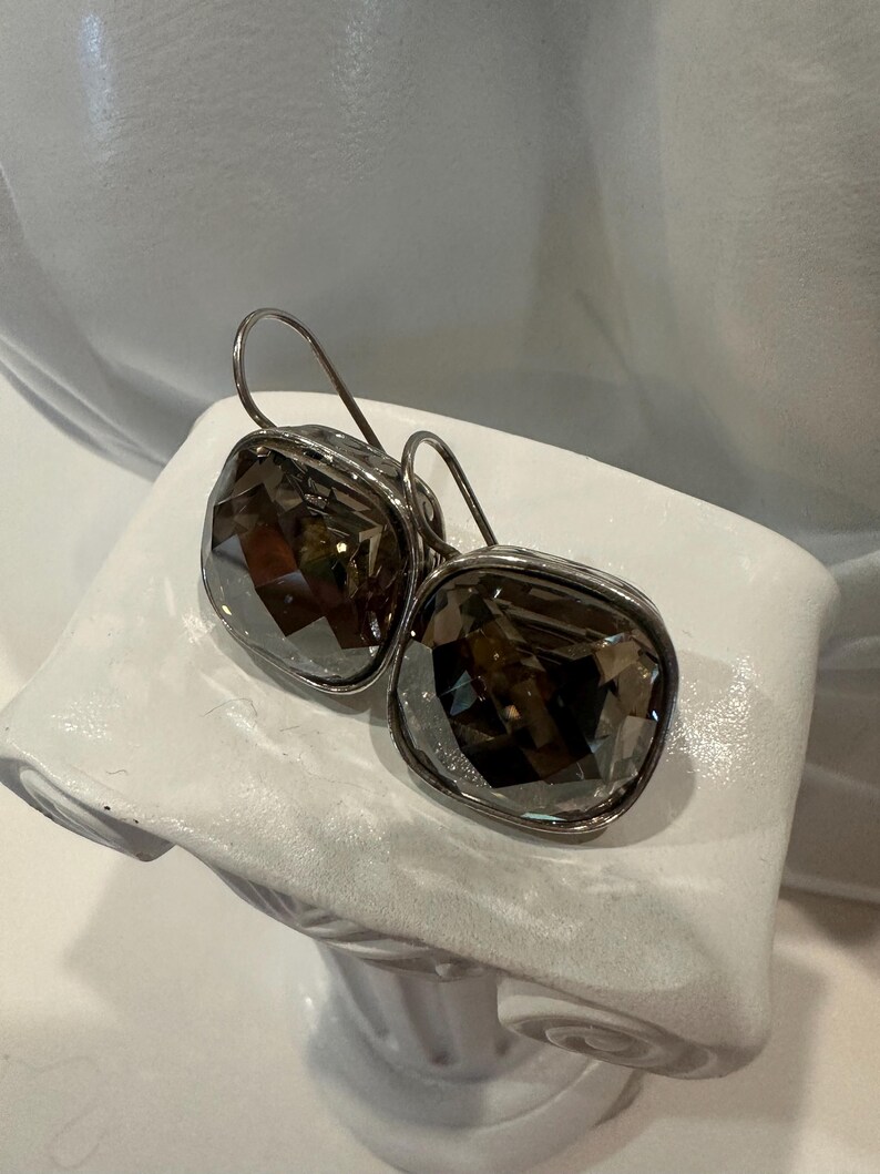 Peut inclure: Une paire de boucles d'oreilles carr&eacute;es en cristal gris fum&eacute; avec des montures en m&eacute;tal argent&eacute; et des fermoirs &agrave; crochet. Les boucles d'oreilles sont pr&eacute;sent&eacute;es sur un buste blanc.