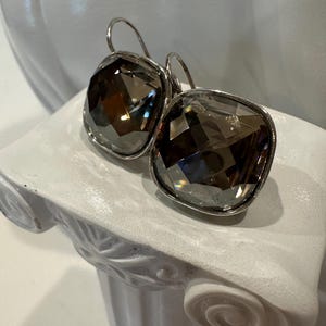 Peut inclure: Boucles d'oreilles pendantes argent&eacute;es avec de grands cristaux carr&eacute;s fum&eacute;s et facett&eacute;s. Les boucles d'oreilles ont un cadre argent&eacute; et une fermeture &agrave; crochet. Elles sont pr&eacute;sent&eacute;es sur un pi&eacute;destal d&eacute;coratif blanc.