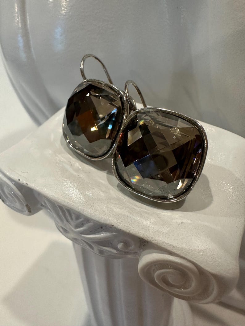 Peut inclure: Boucles d'oreilles pendantes argent&eacute;es avec de grands cristaux carr&eacute;s fum&eacute;s et facett&eacute;s. Les boucles d'oreilles ont un cadre argent&eacute; et une fermeture &agrave; crochet. Elles sont pr&eacute;sent&eacute;es sur un pi&eacute;destal d&eacute;coratif blanc.