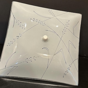 May include: Square, frosted glass ceiling light fixture with a wheat stalk design in black. The fixture has a white, round center piece. The wheat stalks are arranged diagonally across the square. The fixture is on a black surface.