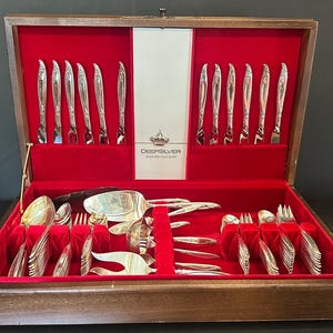 May include: A wooden flatware chest with a red velvet interior containing a set of silver-toned utensils. The center panel features a crown logo and the text DEEPSILVER INLAID WITH SOLID SILVER. The set includes knives, spoons, and forks.