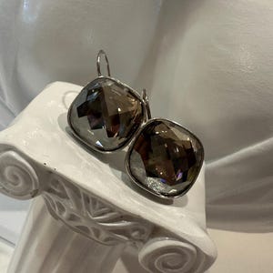 Peut inclure: Une paire de boucles d'oreilles argent&eacute;es avec de grandes pierres carr&eacute;es en quartz fum&eacute; facett&eacute;. Les boucles d'oreilles sont pr&eacute;sent&eacute;es sur un pi&eacute;destal blanc de style classique. Les pierres refl&egrave;tent la lumi&egrave;re.