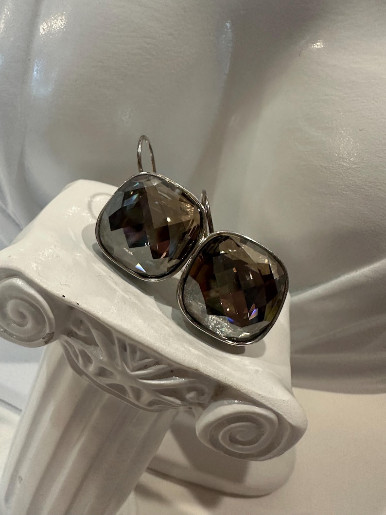 Peut inclure: Une paire de boucles d'oreilles argent&eacute;es avec de grandes pierres carr&eacute;es en quartz fum&eacute; facett&eacute;. Les boucles d'oreilles sont pr&eacute;sent&eacute;es sur un pi&eacute;destal blanc de style classique. Les pierres refl&egrave;tent la lumi&egrave;re.