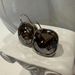 Peut inclure: Une paire de boucles d'oreilles carr&eacute;es en cristal gris fum&eacute; avec des montures en m&eacute;tal argent&eacute;. Les boucles d'oreilles sont pr&eacute;sent&eacute;es sur un pi&eacute;destal d&eacute;coratif blanc. Les cristaux ont de multiples facettes.