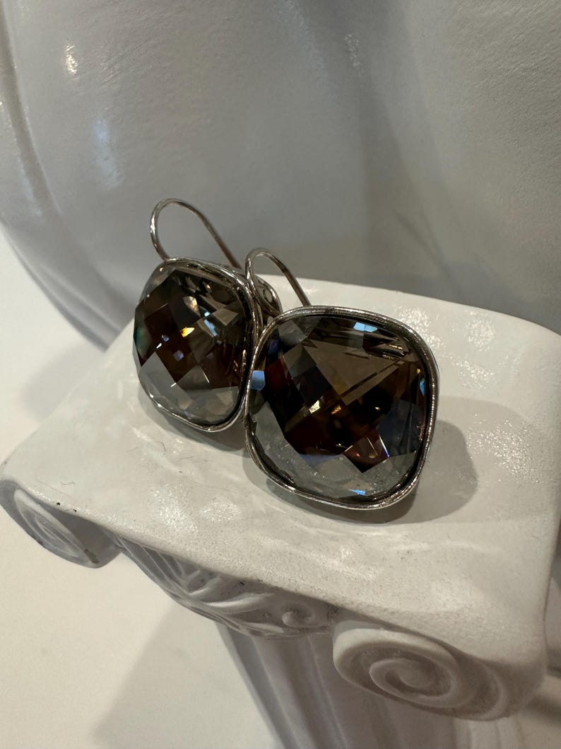 Peut inclure: Une paire de boucles d'oreilles carr&eacute;es en cristal gris fum&eacute; avec des montures en m&eacute;tal argent&eacute;. Les boucles d'oreilles sont pr&eacute;sent&eacute;es sur un pi&eacute;destal d&eacute;coratif blanc. Les cristaux ont de multiples facettes.