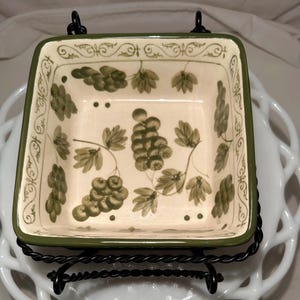 May include: Square ceramic dish with a green border and grape cluster design. The dish is supported by a black metal stand. The grapes are green and brown, with green leaves. The background is off-white.