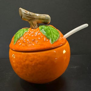 May include: A bright orange ceramic jar shaped like a citrus fruit with a textured surface. The lid features a sculpted brown stem and two green leaves. A white ceramic spoon is inserted into the side of the lid, resting against the orange body.